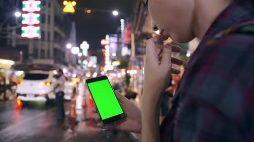 Female tourist holding smart phone with green screen display while standing on the street at night.