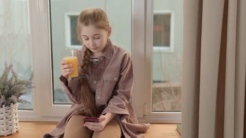 Girl Drinks Juice and Uses Phone by Window