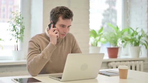 Mann spricht auf dem Smartphone, während er Laptop im Büro benutzt