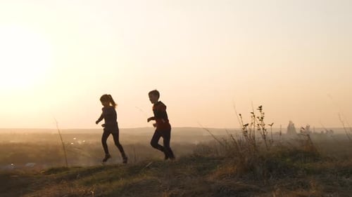 Two children boy and girl running together outdoors on a summer field at sunset.