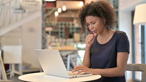 Young African Woman Using Laptop Coughing in Cafe