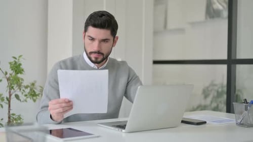 Young Man with Laptop Reading Documents in Office
