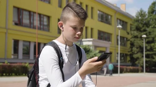A Caucasian Teenage Boy Listens to Music with Earphones on a Smartphone a School in the Background