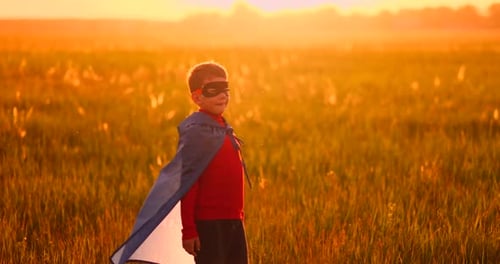 A Child in the Costume of a Superhero in a Red Cloak Runs Across the Green Lawn Against the Backdrop