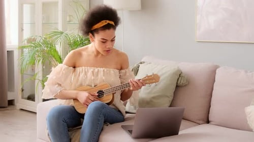 Woman Plays Ukulele on Sofa with Laptop