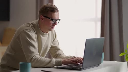 A Man with Glasses Works From Home in His Kitchen Using a Laptop and the Internet