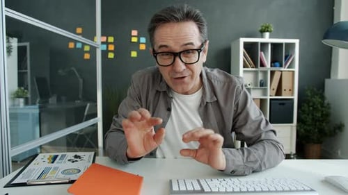 Mature Man in Glasses Speaking and Gesturing Making Internet Video Call in Office at Desk