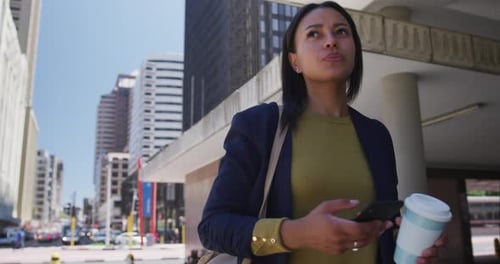 African american woman holding coffee and using smartphone in street