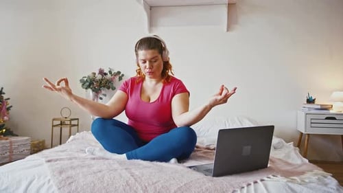 Young Overweight Lady Meditating at Home Sitting on Bed in Headphones and Watching Yoga Lesson on