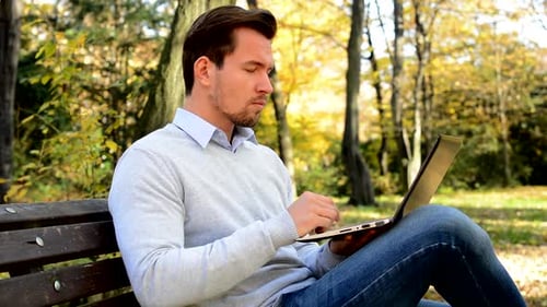Young Handsome Man Sitts on Bench in a Park and Works with a Laptop
