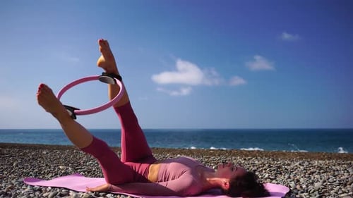A Young Woman with Black Hair Doing Pilates with the Ring on the Yoga Mat Near the Sea on the Pebble
