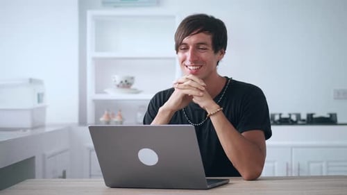 Smiling Man Reading Good News or Watching Funny Videos Using Computer at Home in the Kitchen