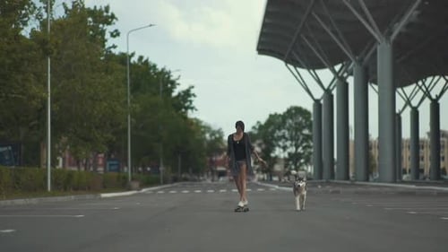 Young Beautiful Woman Riding on Skateboard or Longboard on the Street with Her Husky Dog