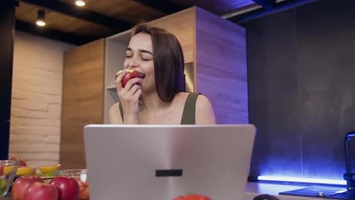 Woman Eats Apple While Using Laptop in Kitchen