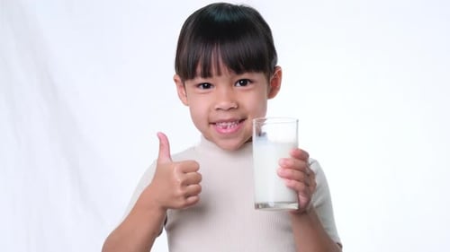 Cute Asian little girl holding a glass of milk and showing thumbs up on white background in studio.