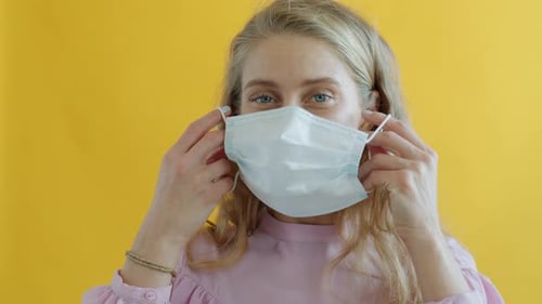 Woman Puts on Surgical Mask on Yellow Background