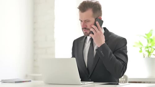 Young Businessman Talking on Phone While Using Laptop in Office