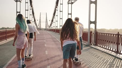 Rear View of Four Young Men Boys and Girls Riding Skateboards Across the Large City Bridge at Dawn
