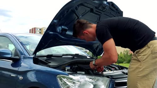 Young Handsome Man Looks and Checks Engine in Car - Buildings in Background