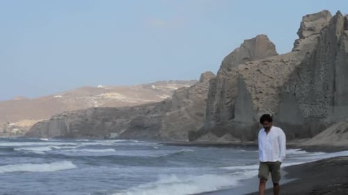 A man is walking all alone at a black sand beach surrounded by white volcanic cliff formations on th