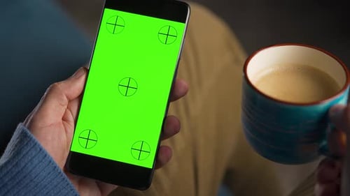 Mann benutzt Smartphone mit grünem Mockup-Bildschirm im vertikalen Modus und trinkt Kaffee