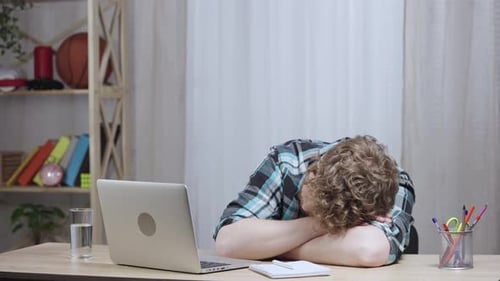 Junger müder Mann im karierten Hemd schläft am Tisch, wacht auf und tippt auf Laptop-Tastatur