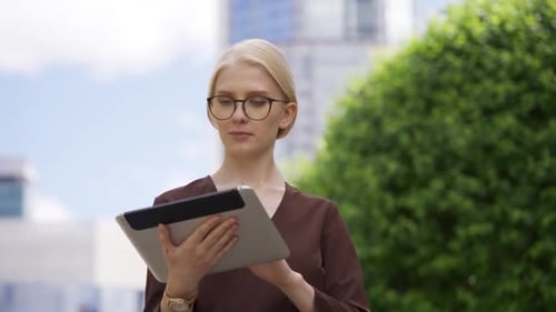 A Young Blonde Woman in Glasses Scrolls the Tablet with Interest in the Street