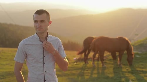 Jovem está parado no campo perto das montanhas. Atrás dele pastando cavalos