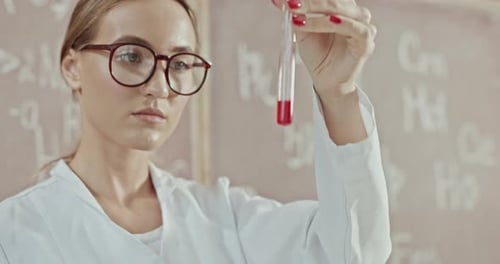 Profesora enfocada en bata de laboratorio y gafas agitando sustancia líquida roja en tubo de ensayo de vidrio