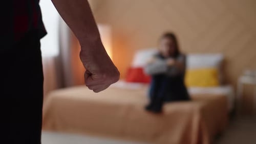 Aggressive man clenching his fist stands in front scared woman sitting on bed, back view, closeup.