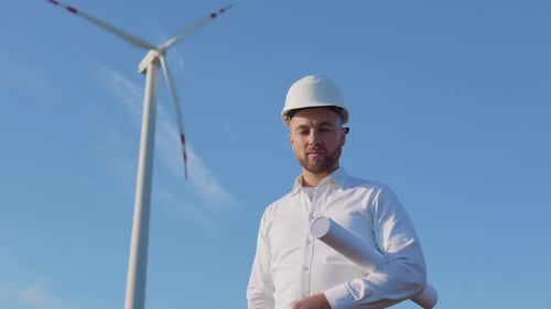 A Male Engineer in a White Helmet and a Classic Shirt Stands Near the Wind Turbines Witha Large