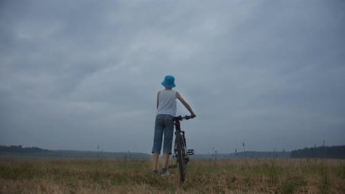 Boy with a Bicycle in a Field in Rainy Weather Looks Around