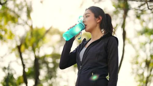 4K Asian woman drinking water from a bottle while jogging at public park in the morning.