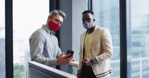 Diverse male office colleagues wearing face masks discussing over smartphone at modern office