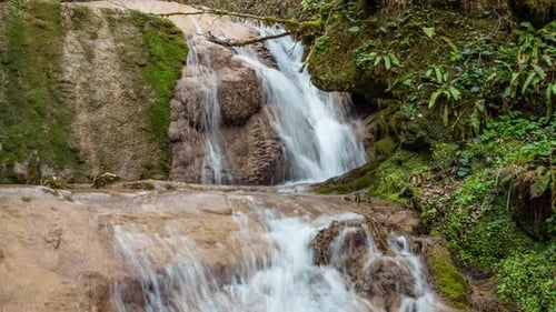 Zoom out view of a waterfall surrounded by moss-covered rocks in a green forest.