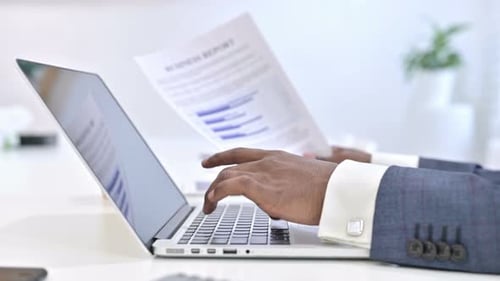 Close Up of Hands of African Man Reading Documents and Working on Laptop