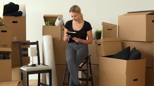 A Moving Woman Sits on a Chair in an Empty Apartment and Writes Into a Notebook, Surrounded By Boxes