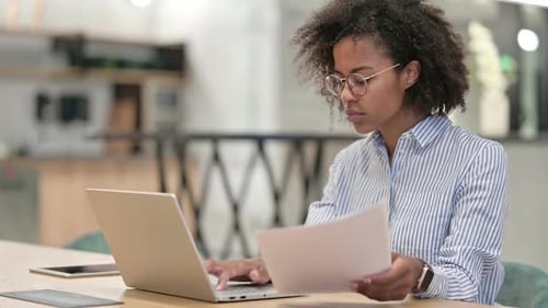 Focused African Businesswoman with Laptop Reading Documents in Office