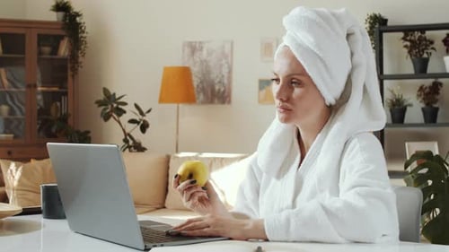 Woman in Bathrobe Works On Laptop While Eating Apple