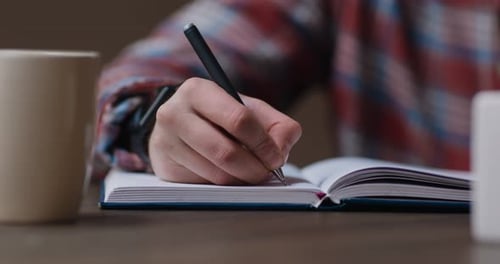 Close Up Shot of Man Writing Plan in Notebook or Filling Diary Sitting at Desk with Coffee at Home