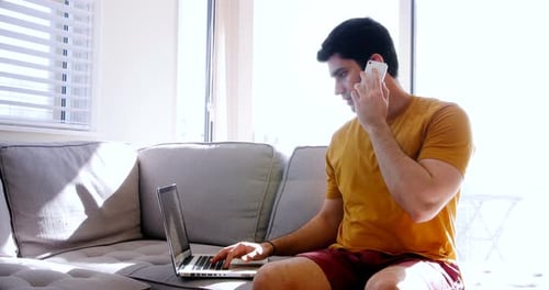 Young Man on Phone, Working on Laptop at Home