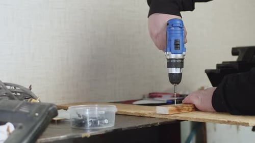 Craftsman works in workshop screwing screw into wooden board with screwdriver, side view. Man screws