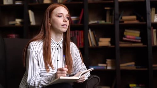 Portrait d'une jolie jeune journaliste aux cheveux roux interrogeant une personne dans une chambre sombre et confortable.