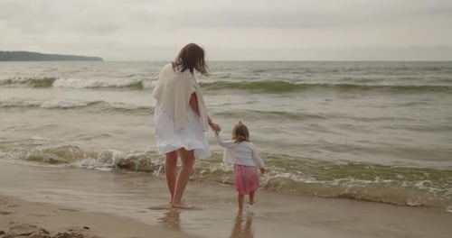 Mother and Baby Daughter Walk on the Beach Near the Sea Together Holding Hands