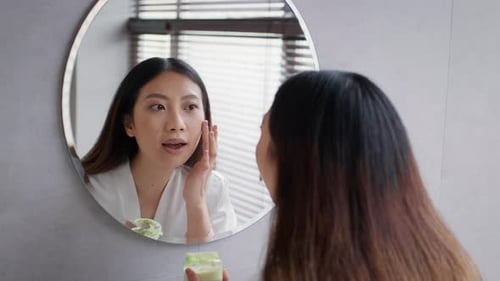 Woman Applies Face Cream in Bathroom Mirror
