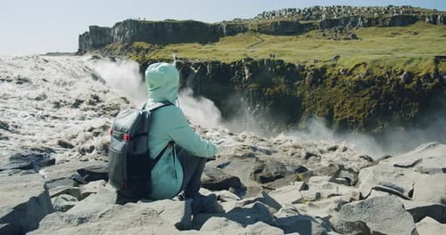 Frau sitzt auf Felsen und genießt den mächtigen Detifoss-Wasserfall und den Basalt Canyon in Island