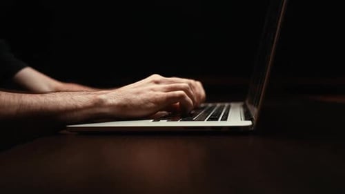 Dollyin-Aufnahme eines jungen Mannes, der in Dunkelraum-Nahaufnahme auf Tastatur und Laptop tippt