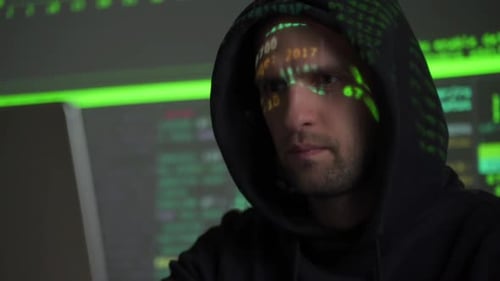 Männlicher Hacker arbeitet an einem Computer, während Green-Code-Charaktere in einem dunklen Büro über sein Gesicht reflektieren