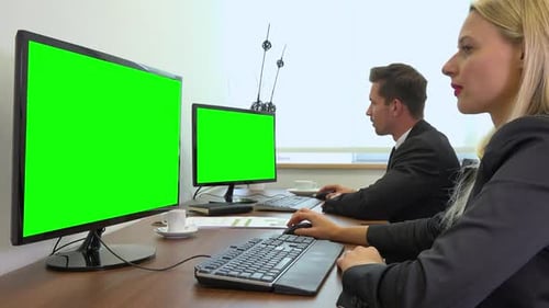 Two office workers, man and woman, work on computers with green screens and talk