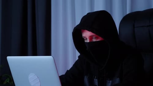 Hacker face in black hood looking at computer and typing codes